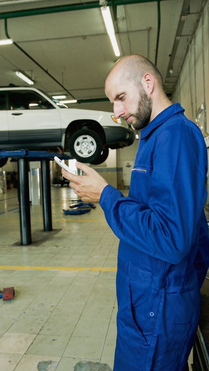 Bald mechanic in blue jumpsuit using phone in an auto repair garage with a car on the lift.