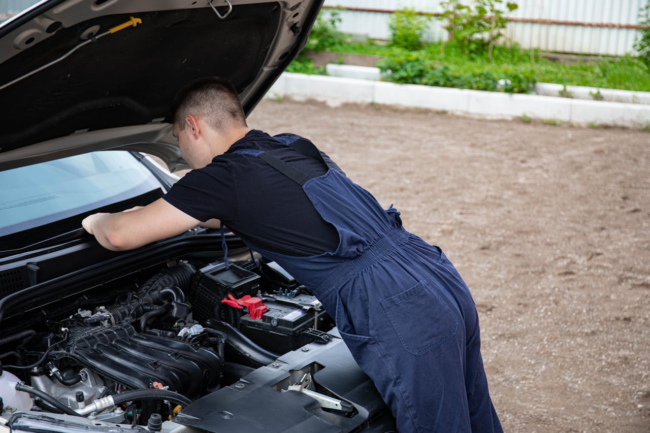 Mechanic in blue coveralls working on a car engine outdoors, focused on repair.